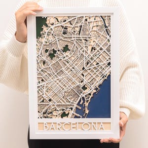 May include: A framed, three-dimensional map of Barcelona, Spain, with a white frame. The map is made of layered wood, showing streets, buildings, and green park areas. The word "BARCELONA" is cut out at the bottom. The map is held by a person.