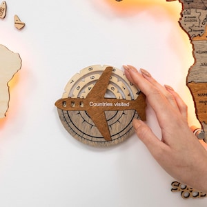 May include: A wooden airplane-shaped travel tracker with the text "Countries Visited" and a compass design. The tracker is being placed on a wooden world map. The airplane has a dark brown finish, and the compass has a light brown finish.