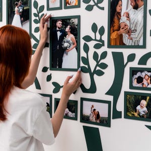 May include: A white wall with a green tree-shaped frame holding various photos. The photos are of a couple, a family, and a baby. The frame is made of wood and has a rustic look.