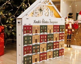 May include: A white wooden advent calendar shaped like a house, with 24 small doors painted to resemble houses. The calendar is labeled "Noah's Winter Surprises" and features a nativity scene cutout at the top. Each door is numbered.
