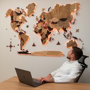 May include: A large, multi-layered wooden world map wall art piece featuring various shades of brown and black wood, illuminated by warm LED backlighting. The map includes a compass rose, small airplane and sailboat icons, and the text ANTARCTICA.