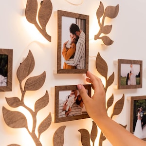 Puede incluir: Una decoraci&oacute;n de pared con la silueta de un &aacute;rbol de madera con hojas y ramas, iluminadas por una cadena de luces. El &aacute;rbol est&aacute; decorado con varios marcos de fotos, cada uno con una fotograf&iacute;a diferente de una familia. Los marcos est&aacute;n dispuestos en un grupo, creando una exhibici&oacute;n cohesiva y conmovedora.