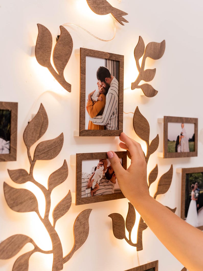 Puede incluir: Una decoraci&oacute;n de pared con la silueta de un &aacute;rbol de madera con hojas y ramas, iluminadas por una cadena de luces. El &aacute;rbol est&aacute; decorado con varios marcos de fotos, cada uno con una fotograf&iacute;a diferente de una familia. Los marcos est&aacute;n dispuestos en un grupo, creando una exhibici&oacute;n cohesiva y conmovedora.