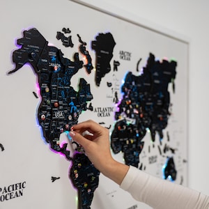 Könnte beinhalten: Eine schwarze Weltkarte mit LED-Leuchten an den Rändern, an einer weißen Wand befestigt. Die Karte zeigt Umrisse von Ländern und Stadtmarkierungen. Eine Hand platziert eine Stecknadel auf dem USA-Abschnitt. Es ist ein dekorativer Artikel.