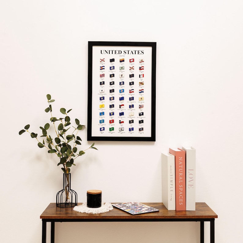 May include: A framed print displaying the flags of the United States. The black framed print is mounted on a white wall above a wooden table. The table holds a vase with greenery, a candle, and books.