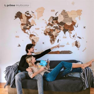 Pu&ograve; includere: Una grande mappa del mondo in legno multicolore montata su una parete bianca. La mappa &egrave; realizzata in varie tonalit&agrave; di legno marrone e beige. Una coppia &egrave; seduta su un divano grigio scuro, con l'uomo che indica la mappa. Il testo "L prime Multicolor" &egrave; in alto a sinistra.