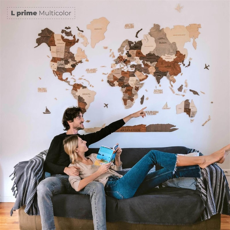 Pu&ograve; includere: Una grande mappa del mondo in legno multicolore montata su una parete bianca. La mappa &egrave; realizzata in varie tonalit&agrave; di legno marrone e beige. Una coppia &egrave; seduta su un divano grigio scuro, con l'uomo che indica la mappa. Il testo "L prime Multicolor" &egrave; in alto a sinistra.