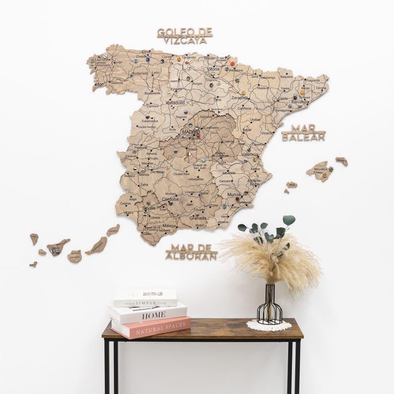 Peut inclure: Une carte en bois d&eacute;taill&eacute;e de l'Espagne, avec des noms de villes et des &eacute;pingles, fix&eacute;e sur un mur blanc. La carte porte les inscriptions "GOLFO DE VIZCAYA", "MAR BALEAR" et "MAR DE ALBORAN". Une table en dessous contient des livres et un vase d&eacute;coratif.