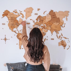 May include: A detailed wooden world map, with country names, is mounted on a white wall. A person is reaching towards the map. A compass rose is in the lower left corner. The map is made of various shades of brown wood.