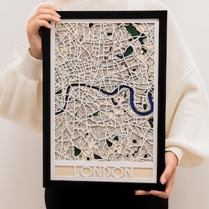 Op de afbeelding: Een ingelijste papieren kaart van Londen, met gedetailleerde straten en groene gebieden, met de rivier de Theems in blauw. De kaart is geplaatst in een zwarte lijst, met de naam van de stad uitgesneden. Een gedetailleerde weergave.