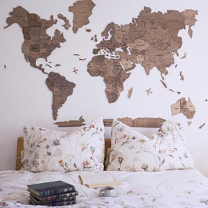 Peut inclure: Une grande carte du monde en bois fixée au mur blanc au-dessus d'un lit. La carte est composée de pièces de bois individuelles, avec les noms des pays visibles. Deux oreillers à motifs floraux sont sur le lit, avec des livres et une tasse.