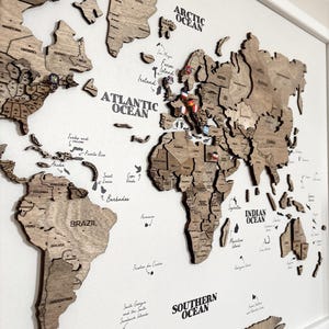 May include: A wooden world map with country outlines and ocean names in a white frame. The map is made of light brown wood and includes pins marking locations. The oceans are labeled in black text. A decorative piece for travel enthusiasts.