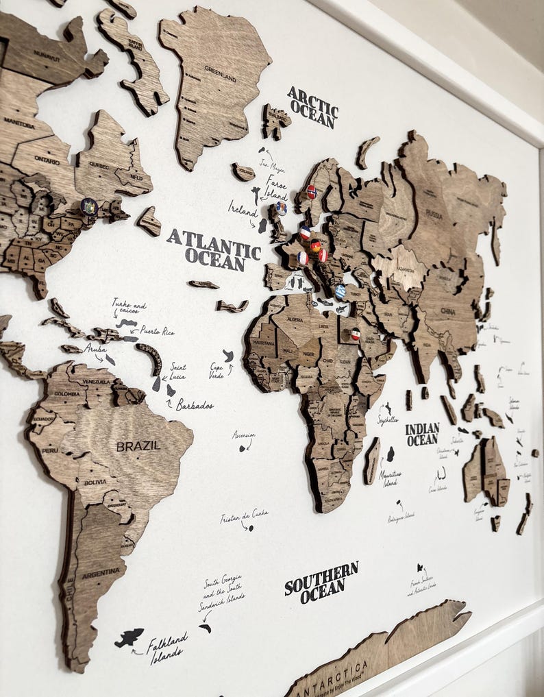 May include: A wooden world map with country outlines and ocean names in a white frame. The map is made of light brown wood and includes pins marking locations. The oceans are labeled in black text. A decorative piece for travel enthusiasts.