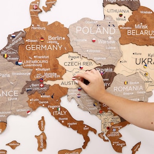 Könnte beinhalten: Eine Holzkarte von Europa mit Ländergrenzen und Städtenamen. Die Karte besteht aus hellem und dunklem braunem Holz. Eine Hand platziert eine kleine Flaggennadel auf Österreich. Die Karte enthält Länderflaggen.