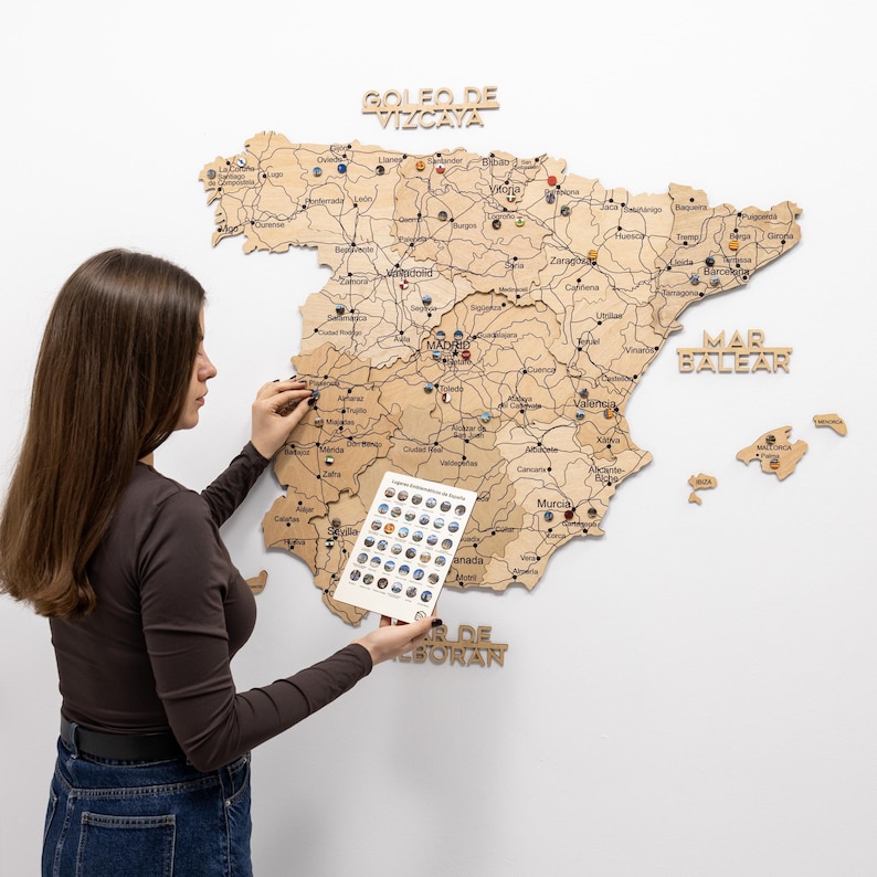 Peut inclure: Une carte en bois de l'Espagne avec des marqueurs de villes et du texte. La carte est fix&eacute;e sur un mur blanc. Une personne tient une carte avec des marqueurs suppl&eacute;mentaires. La carte comprend le texte "Golfo de Vizcaya", "Mar Balear" et "Mar de Alboran".