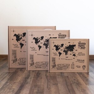 May include: Three rectangular cardboard boxes of varying sizes, each featuring a black printed world map, a compass rose, and illustrations of a living room interior. The text reads Wooden Travel Map, Travel & Mark visited spots, and Decor & Perfect photo zone.