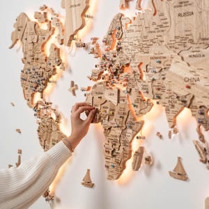 May include: A large, multi-layered wooden world map wall art piece featuring engraved country names, borders, and integrated warm LED backlighting. The map includes small decorative wooden airplane and sailboat icons, with colorful push pins.