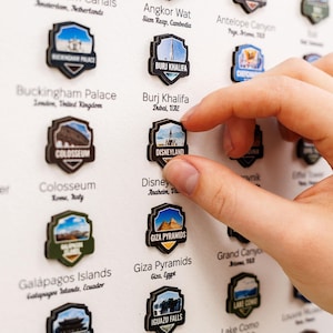 May include: A travel-themed wall decor featuring shield-shaped magnets with iconic landmarks. The magnets include locations like Disneyland, Burj Khalifa, and the Colosseum. The image shows a hand placing a Disneyland magnet on the board.