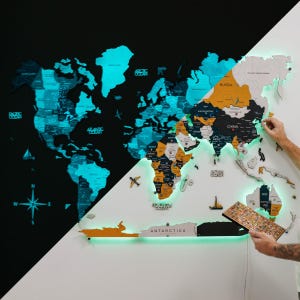 Pode incluir: Um grande mapa-múndi de madeira iluminado, com um fundo preto e branco. O mapa apresenta países em várias cores, com uma borda verde brilhante e oceanos turquesa. Uma pessoa está segurando uma pequena peça de madeira, adicionando-a ao mapa.