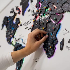May include: A black world map with LED lights outlining the continents. A hand is placing a pin on the map. The map features country names and pin locations. The map is mounted on a white wall.