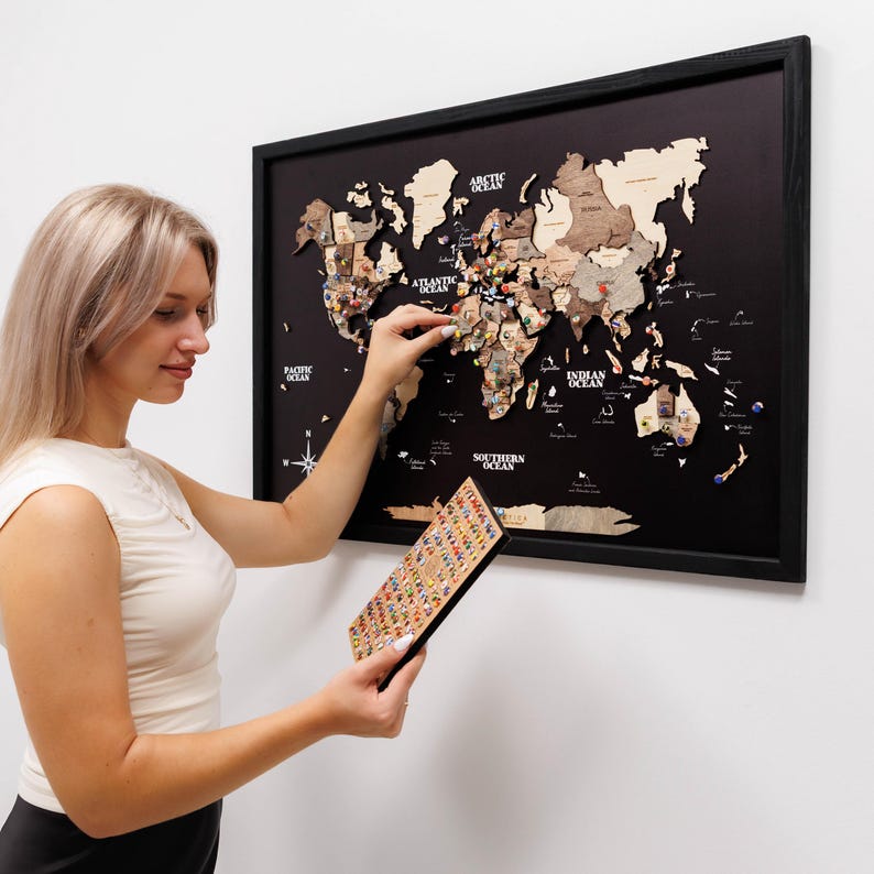 May include: A framed wooden world map with a black background and wooden continents. The map is decorated with colorful push pins. A person is holding a small board of push pins and placing a pin on the map.