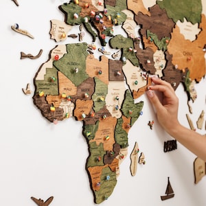 May include: A multi-layered wooden world map wall art featuring various shades of brown and green wood tones. The map includes laser-cut country names and is decorated with numerous small push pins featuring various national flag designs.