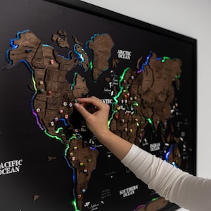 May include: A wooden world map with colourful LED lights outlining the continents. The map is mounted on a black background and framed. A person is placing a pin on the map. The map includes the text "PACIFIC OCEAN", "ARCTIC OCEAN", and "SOUTHERN OCEAN".