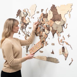 May include: A large, multi-layered wooden world map wall art piece featuring various shades of brown and beige wood. The map includes laser-cut continents, ocean names, and decorative wooden icons like ships and planes, with colorful pins inserted.