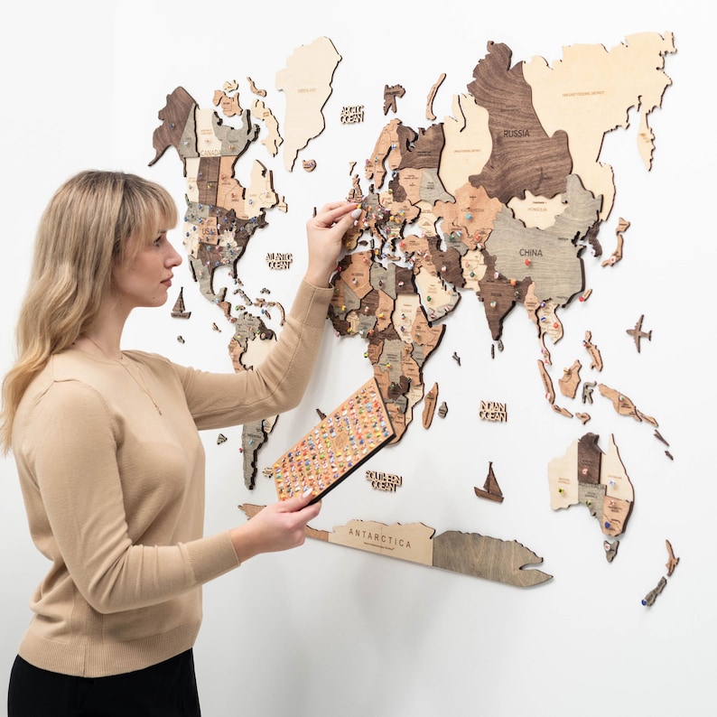 May include: A large, multi-layered wooden world map wall art piece featuring various shades of brown and beige wood. The map includes laser-cut continents, ocean names, and decorative wooden icons like ships and planes, with colorful pins inserted.