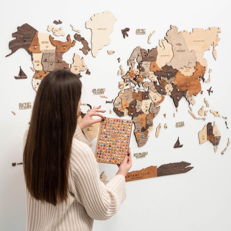 May include: A large wooden world map wall art installation featuring various shades of brown and tan wood pieces. The map includes labels for continents and oceans, with small decorative wooden icons like ships and planes. A rectangular board holds many colorful pins.