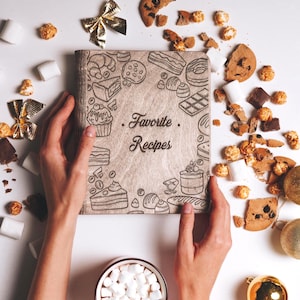 K&ouml;nnte beinhalten: Ein Rezeptbuch aus Holz mit braunem Einband und wei&szlig;em Hintergrund. Der Einband zeigt eine schwarz-wei&szlig;e Illustration verschiedener Desserts und den Text "Favorite Recipes".