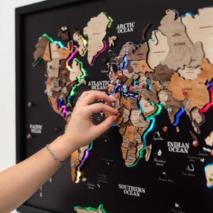Peut inclure: Une carte du monde en bois encadrée de noir, avec des lumières LED colorées soulignant les continents. La carte est ornée de découpes de pays en bois et d'épingles. Une main place une épingle sur la carte.