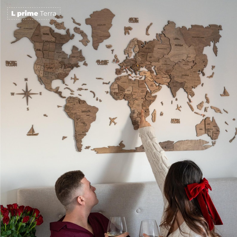 May include: A large wooden world map wall art piece featuring laser-cut continents and countries in varying shades of brown wood. The map includes text labels for major oceans and countries, along with decorative wooden icons of airplanes and sailboats.