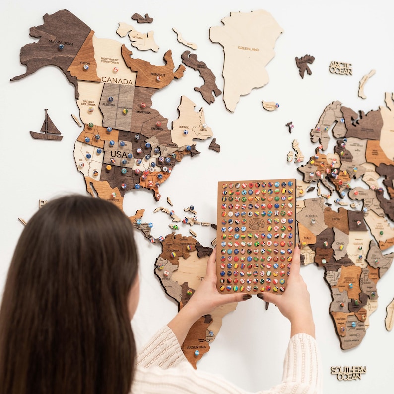 May include: A wooden world map with colourful push pins marking locations. The map is mounted on a white wall. A person is holding a small book with a grid of colourful flags.