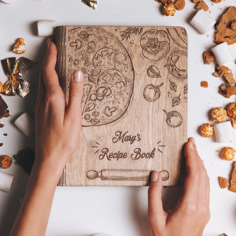 K&ouml;nnte beinhalten: Ein Rezeptbuch aus Holz mit einem braunen Holzdeckel. Der Deckel ist mit einem eingravierten Design einer Pizza, Gem&uuml;se und einem Nudelholz verziert. Der Text "Mary's Recipe Book" ist in den Deckel graviert.