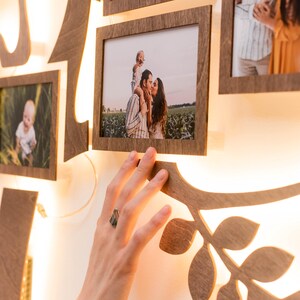 May include: A wooden family tree photo display with a warm white light behind each frame. The frames are arranged in a tree shape and feature photos of a family, including a couple kissing, a young child being held by a parent, and a baby.