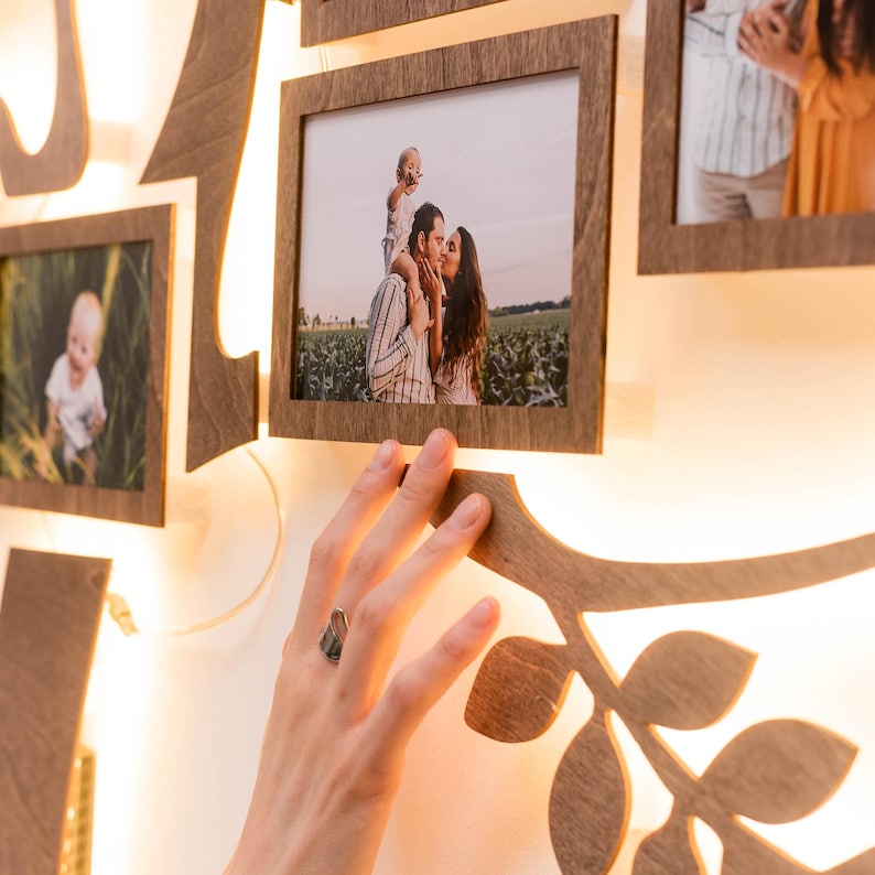 May include: A wooden family tree photo display with a warm white light behind each frame. The frames are arranged in a tree shape and feature photos of a family, including a couple kissing, a young child being held by a parent, and a baby.