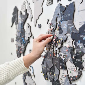 May include: A multi-layered wooden world map wall art piece featuring dark gray and black continents with white text labels for countries and regions. The map is decorated with numerous small, colorful flag-themed push pins placed across various locations.