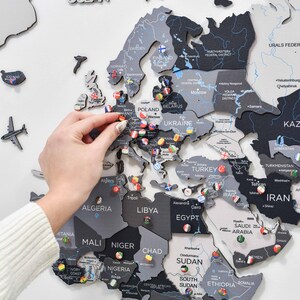 May include: A wall-mounted wooden world map featuring a monochromatic color palette of black, dark gray, and light gray. The map includes laser-cut country names and borders, decorated with small circular pins displaying various national flags.