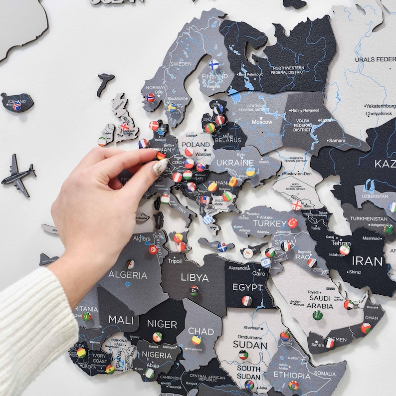 May include: A wall-mounted wooden world map featuring a monochromatic color palette of black, dark gray, and light gray. The map includes laser-cut country names and borders, decorated with small circular pins displaying various national flags.