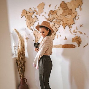 Pu&ograve; includere: Una donna con un cappello marrone e una macchina fotografica &egrave; in piedi davanti a una mappa del mondo in legno su un muro bianco. La mappa &egrave; composta da singoli pezzi di legno, con i nomi dei paesi incisi. La donna indossa un top fantasia e pantaloni scuri.