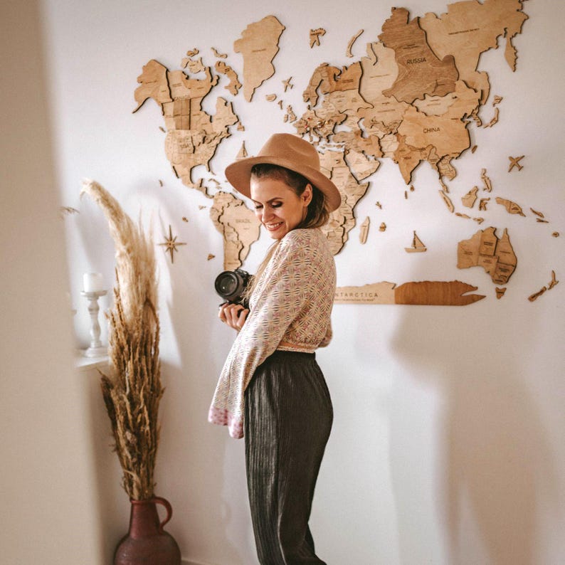 Pu&ograve; includere: Una donna con un cappello marrone e una macchina fotografica &egrave; in piedi davanti a una mappa del mondo in legno su un muro bianco. La mappa &egrave; composta da singoli pezzi di legno, con i nomi dei paesi incisi. La donna indossa un top fantasia e pantaloni scuri.