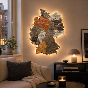 Op de afbeelding: Een wandkaart van hout van Duitsland met afzonderlijke deelstaten in tinten bruin, grijs en wit. De kaart bevat gegraveerde stadsnamen en labels van deelstaten, verlicht van achteren door een warm LED-licht dat een zachte gloed creëert.