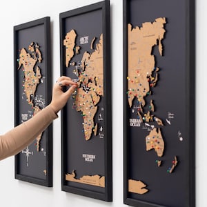 Può includere: Tre mappe da parete rettangolari verticali con continenti in legno tagliati al laser su sfondi neri all'interno di cornici nere. Le mappe mostrano etichette geografiche e sono decorate con numerose puntine colorate per segnare varie posizioni.