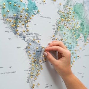 May include: A world map with a teal and white color scheme, dotted with numerous gold push pins. The map shows the continents and oceans, with the words "United States" and "North Atlantic Ocean" visible. A hand is placing a gold pin on the map.