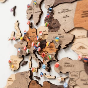 Könnte beinhalten: Eine hölzerne Wandkarte von Europa mit lasergeschnittenen Ländergrenzen und eingravierten Namen von Ländern und Städten. Kleine, runde Stecknadeln mit verschiedenen Nationalflaggen sind in die Holzoberfläche gesteckt, um Orte zu markieren.
