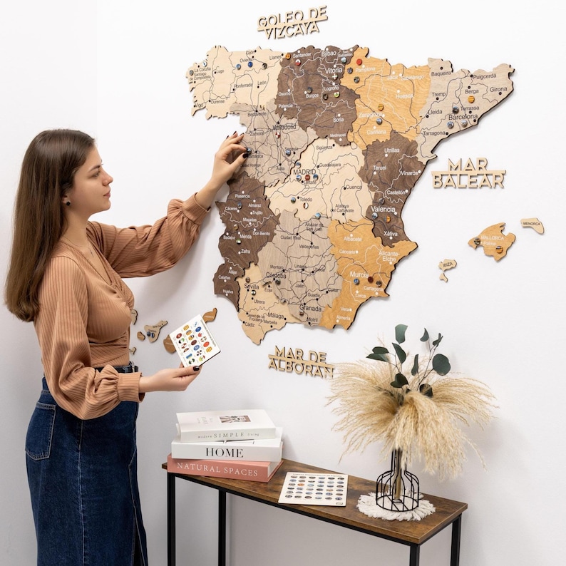 Peut inclure: Une carte en bois de l'Espagne, dans des tons de marron et de beige, fix&eacute;e &agrave; un mur blanc. La carte est &eacute;tiquet&eacute;e avec des caract&eacute;ristiques g&eacute;ographiques et des noms de villes. Une personne place une &eacute;pingle sur la carte. Des pi&egrave;ces de carte et des &eacute;pingles suppl&eacute;mentaires sont sur une table voisine.