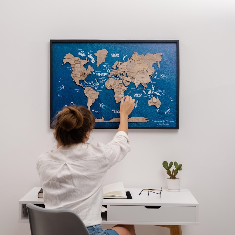 May include: A framed wooden world map with a blue ocean background. The map is detailed with continents in a natural wood tone. The map is hanging on a white wall. A person is pointing at the map.