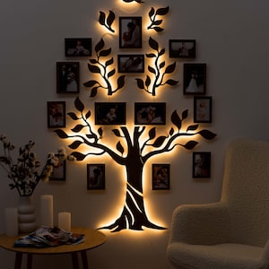 Puede incluir: Una silueta negra de un árbol con ramas y hojas, iluminada con luces LED blancas cálidas. El árbol está decorado con múltiples marcos de fotos negros, cada uno con una fotografía diferente. Una silueta de pájaro está posada en una rama en la parte superior del árbol.
