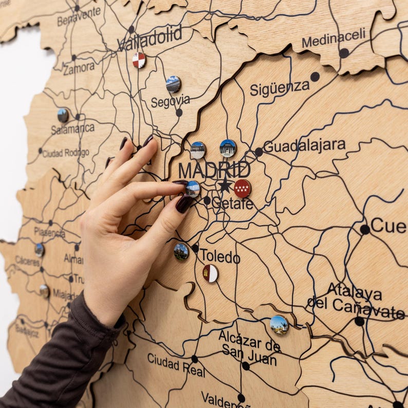 Peut inclure: Carte en bois de l'Espagne avec les noms des villes et les routes grav&eacute;es. De petites &eacute;pingles color&eacute;es marquent les lieux. Une main place une &eacute;pingle sur Madrid. La carte est en bois clair.